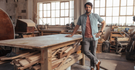 Porträt eines selbstbewussten jungen Holzarbeiters, der in seiner Werkstatt neben einer Holzbank steht
